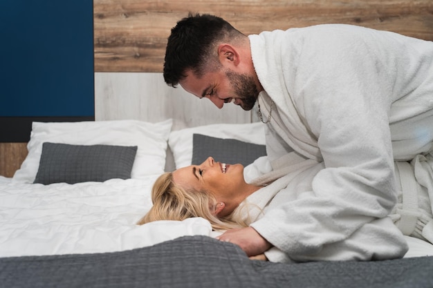 Una pareja joven, un hombre y una mujer con túnicas de spa blancas, se están divirtiendo en una cama en un hotel.