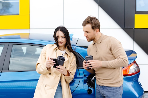 Pareja joven hombre y mujer viajando en automóvil eléctrico con parada en la estación de carga Novio conectando el cable para cargar Hombre hablando con novia sosteniendo una taza bebiendo café caliente sonriendo