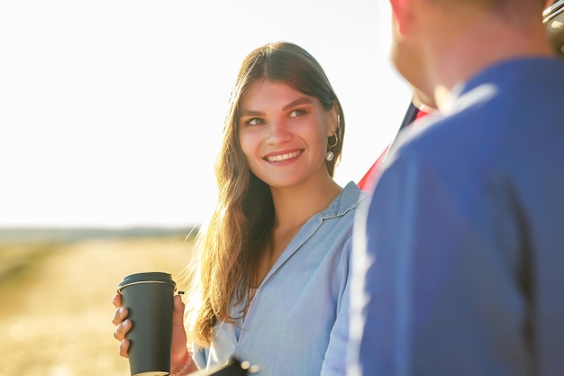 Una pareja joven, un hombre y una mujer, viajando juntos en un auto nuevo con una parada para tomar café en un campo de trigo al atardecer