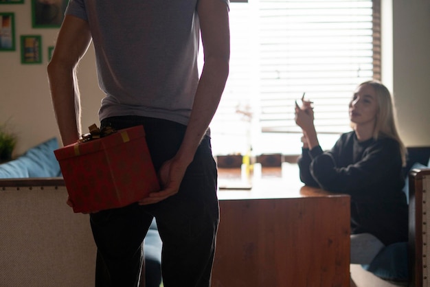 Una pareja joven, hombre sosteniendo una caja de regalo detrás de la espalda, sorprendiendo a su novia