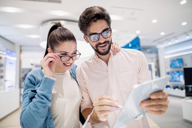 Pareja joven impresionada probando nueva tableta mientras está de pie en la tienda de tecnología.