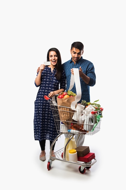 Pareja joven india con carrito de compras o carretilla llena de comestibles, verduras y frutas. Foto de cuerpo entero aislado sobre pared blanca