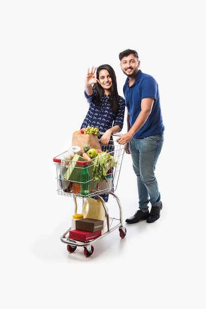 Pareja joven india con carrito de compras o carretilla llena de comestibles, verduras y frutas. Foto de cuerpo entero aislado sobre pared blanca