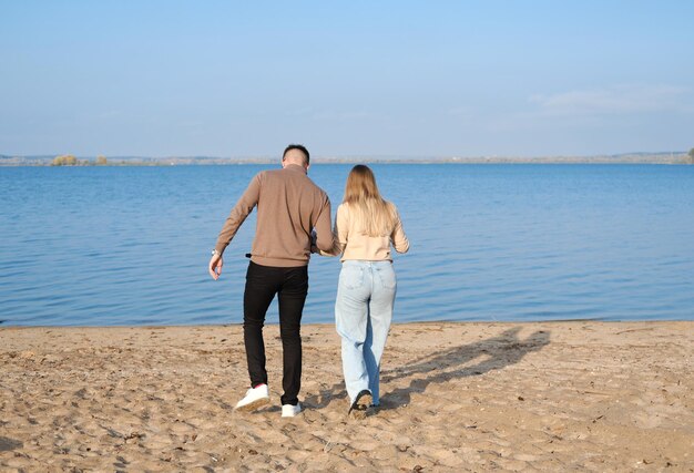 Una pareja joven en jeans y suéteres se toman de la mano y caminan por la playa Vista trasera Clima soleado
