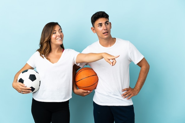 Pareja joven jugando al fútbol y baloncesto en azul apuntando hacia el lado para presentar un producto