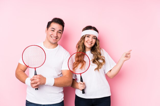 Pareja joven jugando bádminton aislado sonriendo y señalando a un lado, mostrando algo en el espacio en blanco.