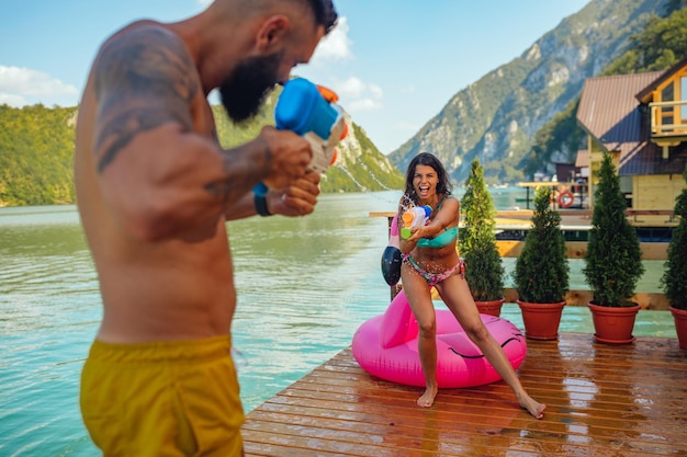 Pareja joven jugando con pistolas de agua en el muelle del lago