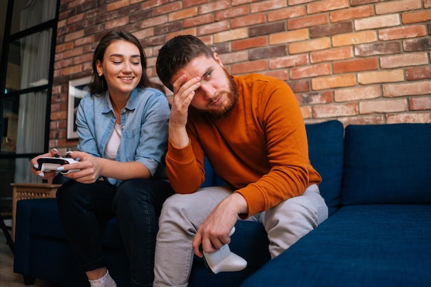 Pareja joven jugando videojuegos en la consola sentados juntos en el sofá en la acogedora sala de estar Señora feliz de ganar la victoria chico sintiéndose decepcionado