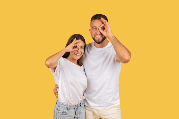 Foto una pareja joven y juguetona con camisetas blancas haciendo gestos divertidos con las manos sobre un ojo