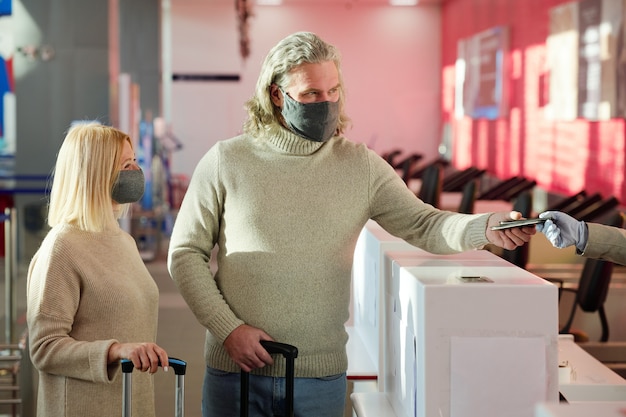 Foto pareja joven en máscaras protectoras de pie con equipaje en el mostrador de recepción y hacer el check-in antes del vuelo