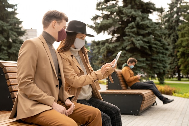 Pareja joven con máscaras protectoras y ropa casual elegante descansando en un banco en el parque mientras mira la pantalla de la tableta y ve videos en línea