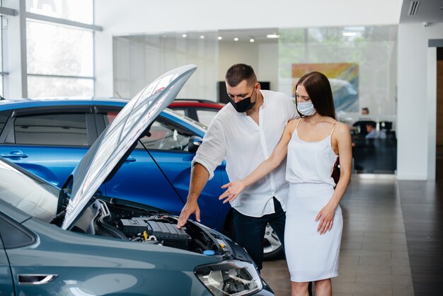 Pareja joven con máscaras selecciona un nuevo vehículo y consulta con un representante del concesionario en el período de la pandemia. Venta de coches y vida durante la pandemia.