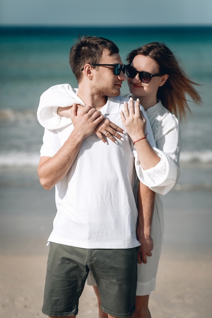 Pareja joven de moda activa en ropa blanca está de pie en la playa y cogidos de la mano. Hermosa rubia con hilo rojo en el brazo y un hombre están de vacaciones. Phuket. Tailandia