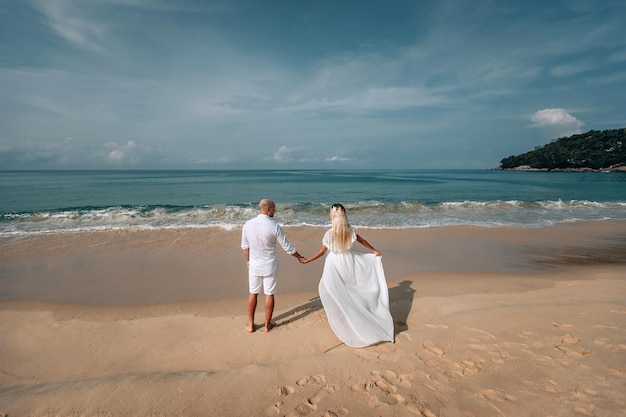 Pareja joven de moda atractiva en ropa blanca está caminando por la playa y tomados de la mano. Hermosa rubia de pelo largo y un hombre calvo están de vacaciones. Phuket. Tailandia