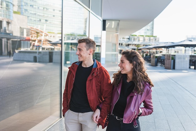 Una pareja joven de moda camina por la ciudad para ir de compras, está feliz por lo que ve en el concepto de amor de la ventana de la tienda Compras navideñas