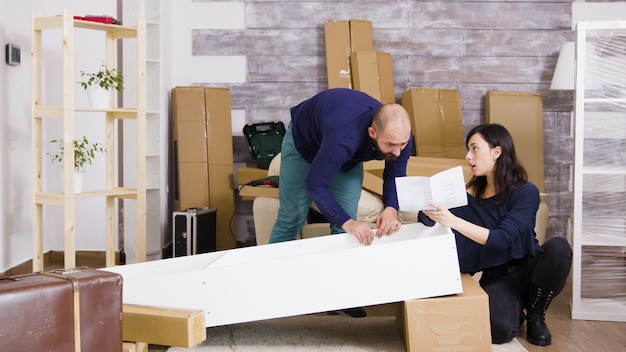 Pareja joven montaje de muebles en equipo en apartamento nuevo. Niña leyendo instrucciones de montaje.