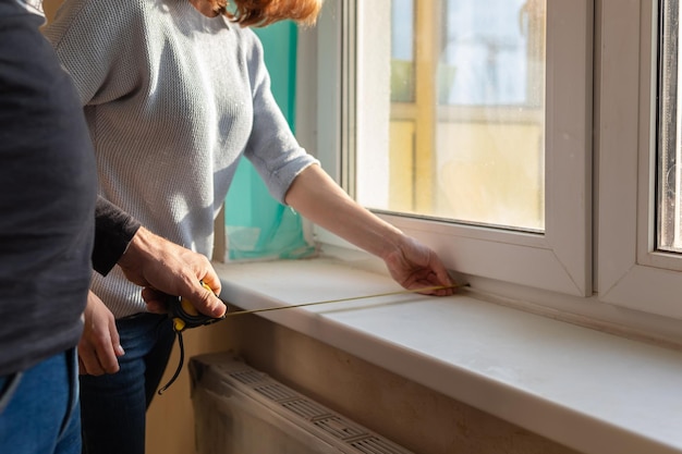 Pareja joven mudándose a un nuevo apartamento La gente mide el tamaño del alféizar de la ventana con una cinta métrica Concepto nuevos hogares nuevo comienzo