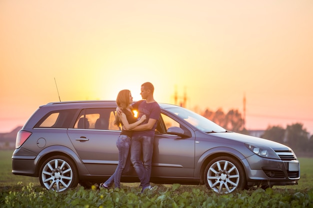 Pareja joven, mujer delgada y atractiva con cabello largo y apuesto hombre deportivo de pie abrazados juntos en el coche plateado en una cálida noche de verano en el cielo brillante al atardecer o amanecer copia espacio de fondo.