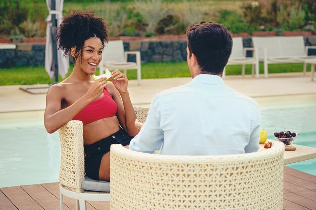 Foto pareja joven multicultural sentado junto a la piscina en una mesa - jóvenes románticos comiendo fruta para el desayuno junto a la piscina del resort - estilo de vida saludable y concepto de viaje