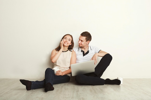 Pareja joven navegando por la web en la computadora portátil sentada en el piso en la sala de estar vacía del nuevo apartamento sobre fondo blanco, mujer hablando por teléfono móvil, espacio de copia