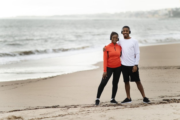 Pareja joven negra positiva en ropa deportiva y abrazos de auriculares inalámbricos relajarse después de correr en el mar