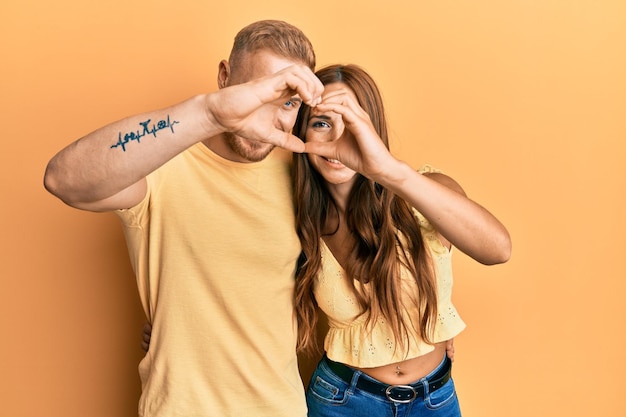 Pareja joven de novia y novio abrazándose y de pie juntos haciendo forma de corazón con la mano y los dedos sonriendo mirando a través de la señal