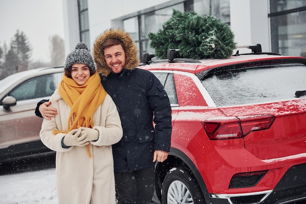 Una pareja joven parada cerca de un auto con un árbol en la parte superior. Juntos al aire libre en invierno.