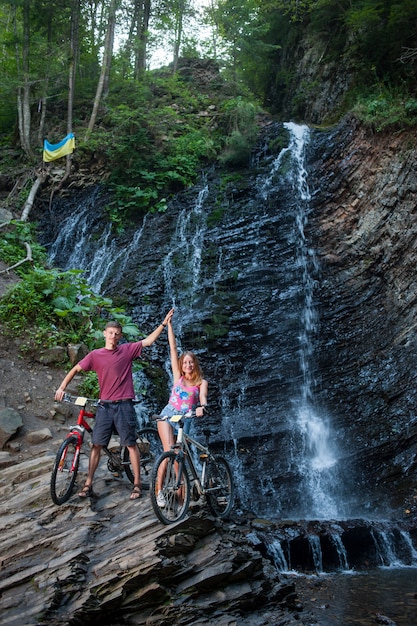 Pareja joven de pie junto a la cascada con sus bicicletas