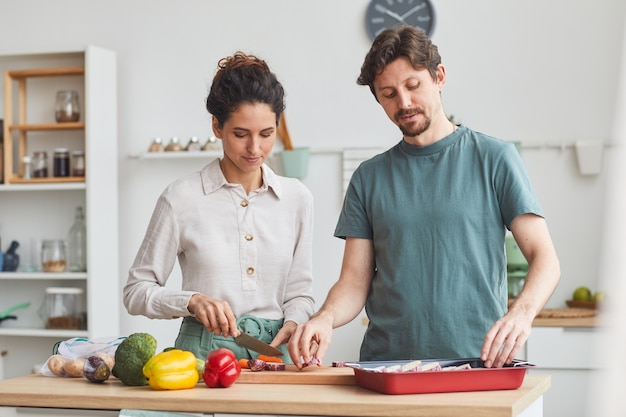 Pareja joven de pie en la mesa de la cocina y preparando la cena juntos en la cocina de casa