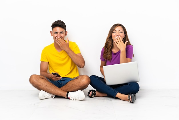 Pareja joven con un portátil sentado en el suelo en blanco feliz y sonriente cubriendo la boca con la mano