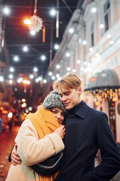 Pareja joven positiva en ropa de abrigo abrazándose en la calle decorada de Navidad.