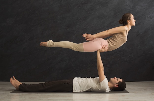 Pareja joven practicando acroyoga en colchoneta en el gimnasio juntos. Mujer volando, copie el espacio, vista lateral. Socio de yoga, flexibilidad, concepto de confianza