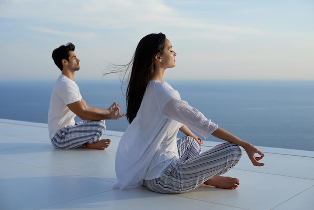 pareja joven practicando yoga al atardecer en una moderna terraza casera con océano y puesta de sol en el fondo