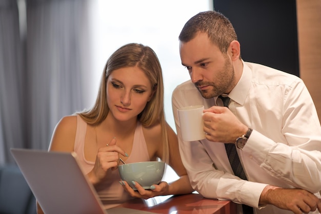 Una pareja joven se prepara para el trabajo y usa una computadora portátil. El hombre bebe café mientras la mujer desayuna juntos en una casa de lujo, mirando la pantalla, sonriendo.