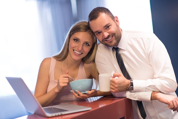 Una pareja joven se prepara para el trabajo y usa una computadora portátil. El hombre bebe café mientras la mujer desayuna juntos en una casa de lujo, mirando la pantalla, sonriendo.