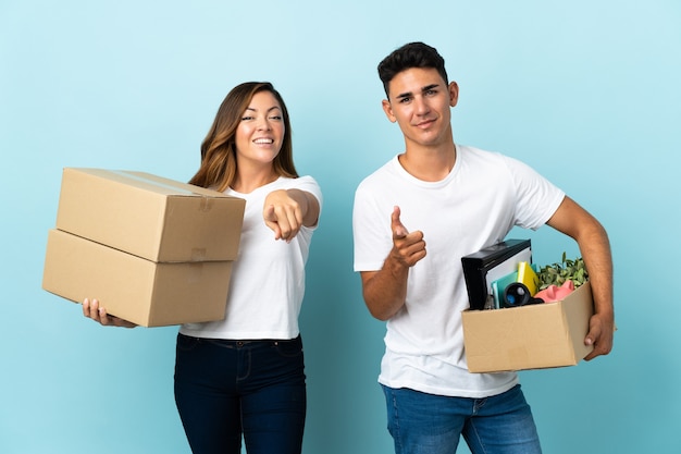 Pareja joven que se muda a una nueva casa entre cajas en azul le señala con el dedo con una expresión de confianza