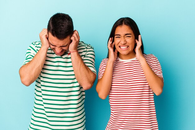 Foto pareja joven de raza mixta aislado en azul cubriendo las orejas con las manos.