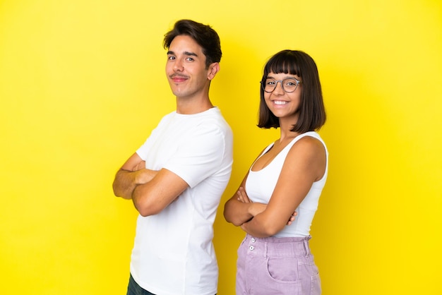 Pareja joven de raza mixta aislado sobre fondo amarillo manteniendo los brazos cruzados en posición lateral mientras sonríe