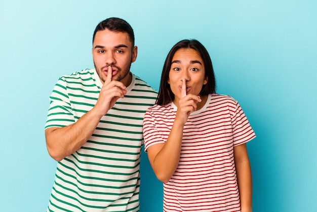 Pareja joven de raza mixta aislado sobre fondo azul manteniendo un secreto o pidiendo silencio.