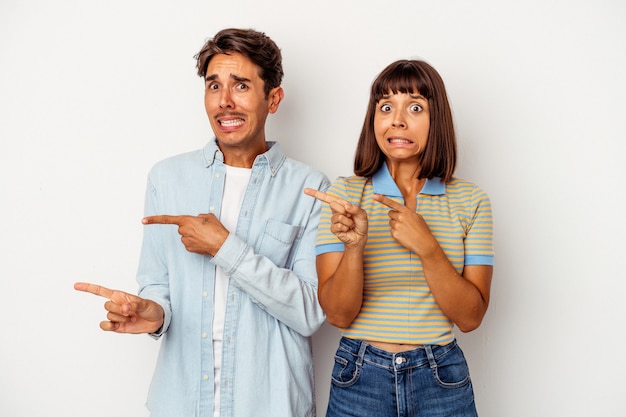 Pareja joven de raza mixta aislado sobre fondo blanco sorprendido apuntando con los dedos índices a un espacio de copia.