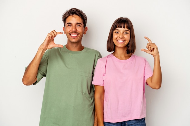 Pareja joven de raza mixta aislado sobre fondo blanco sosteniendo algo poco con los dedos índices, sonriendo y confiado.