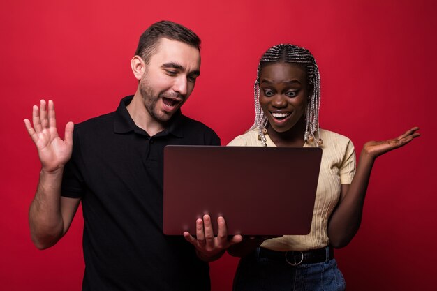 Pareja joven de raza mixta hombre caucásico y mujer africana hoding y usando laptop que se encuentran aisladas sobre fondo rojo.