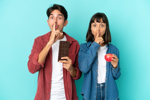 Pareja joven de raza mixta sosteniendo manzana y chocolate aislado sobre fondo azul mostrando un gesto de silencio poniendo el dedo en la boca