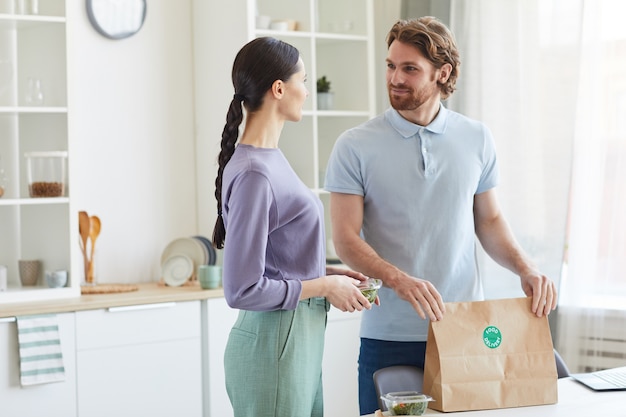 Pareja joven recibiendo comida a domicilio ellos de pie en la cocina y hablando entre sí