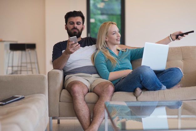 Una pareja joven se relaja en el sofá de la lujosa sala de estar, usando una computadora portátil y un control remoto