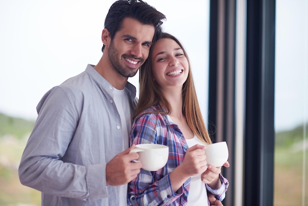 Una pareja joven y relajada bebe el primer café de la mañana sobre una gran ventana luminosa en el interior de una casa moderna