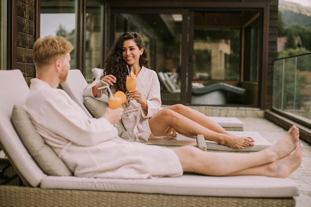 Una pareja joven relajándose en las camas y bebiendo jugo de naranja fresco en la terraza al aire libre