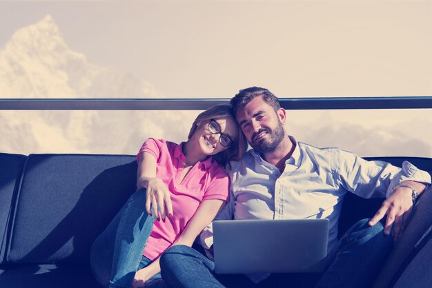 Pareja joven relajándose en casa usando computadoras portátiles leyendo en la sala de estar cerca de la ventana en el sofá cama.