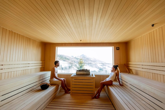 Pareja joven relajándose en la sauna y viendo el bosque de invierno a través de la ventana