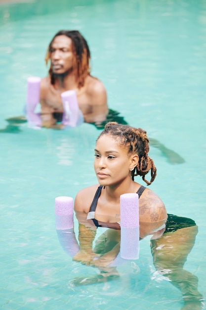 Pareja joven repitiendo después del entrenador al hacer ejercicios con fideos flotantes en la piscina al aire libre del hotel spa